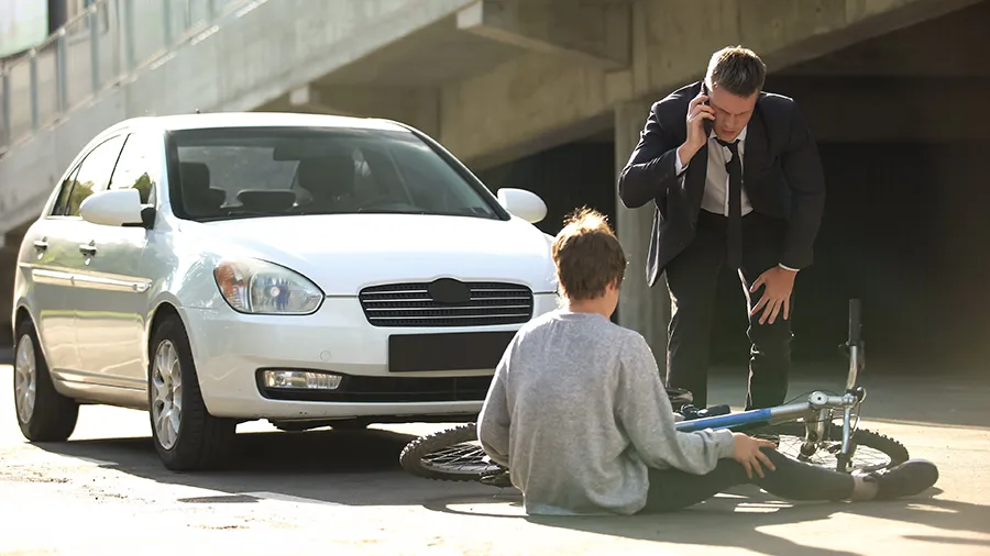 A young man is on the ground in front of a car after bumping his bicycle.